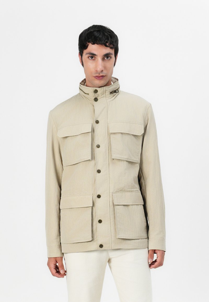 Veste beige avec un col montant, plusieurs poches avant, des fermetures à pression et un tissu texturé. Fini lisse avec un design décontracté.