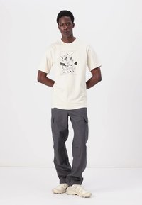 Cremefarvet bomulds t-shirt med et sort grafisk tryk på forsiden, parret med mørkegrå, løstsiddende cargo-bukser. Skoene er beige sneakers.