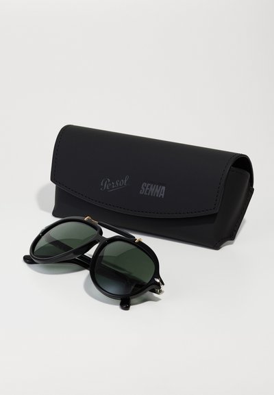 Črne sončna očala Persol z zelenimi lečami, ovalnimi okvirji in kovinskimi detajli, poleg črne usnjene etuija z vtisnjenimi napisi "Persol" in "Senna."