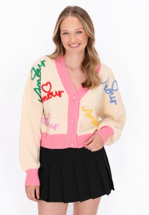 Breede gebreide cardigan in crème met roze accenten, voorzien van meerkleurige geborduurde tekst. Lange, puffy mouwen en ronde knopen aan de voorkant.
