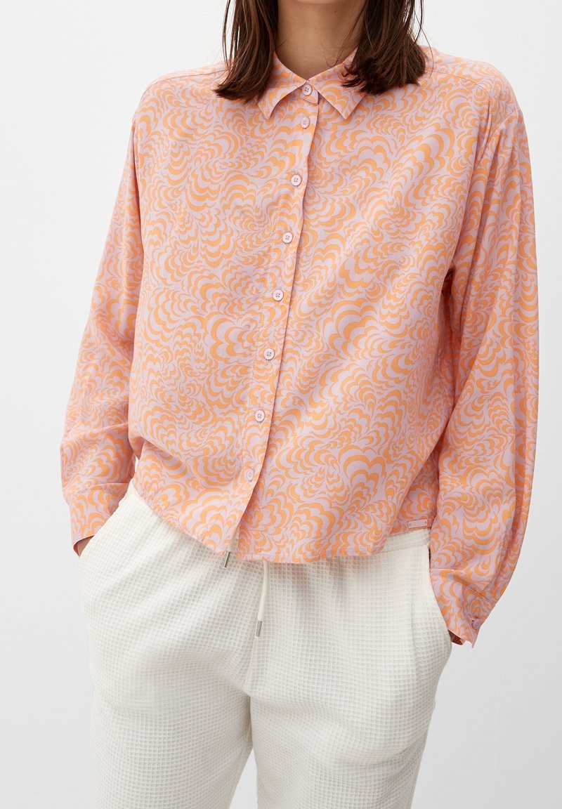 Femme portant une chemise à manches longues boutonnée avec un motif tourbillonnant orange et rose et un pantalon blanc texturé avec des poches à cordon.