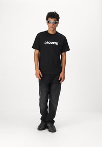 Juodas medvilninis marškinėlis su baltu "LACOSTE" logotipu, derinamas su tamsiomis džinsinėmis kelnėmis ir juodais sportbačiais. Modelis dėvi pilkus akinius nuo saulės.