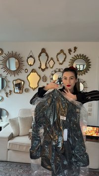 Zwarte, glanzende jas in plastic verpakking, met een klassiek ontwerp met lange mouwen en een puntige kraag. De achtergrond bevat decoratieve spiegels.