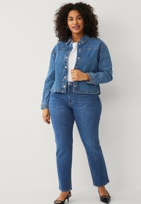 Giacca di jeans blu con colletto classico, chiusura a bottoni e tasche frontali. Indossata sopra una maglietta bianca e abbinata a jeans blu e scarpe nere.
