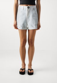 Lys blå denimshorts med oppbrettede kanter, høy livhøyde, frontlommer og svart knappefeste. Brukt med svarte platåflip-flops.
