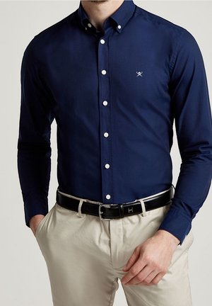 Mann trägt ein marineblaues Hemd mit Knöpfen, kombiniert mit beigen Hosen und einem schwarzen Leder Gürtel mit quadratischer silberner Schnalle, Hände in den Taschen.