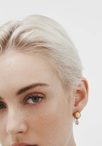 Pendientes deStud en tono dorado con forma redondeada, que presentan un acento en forma de lágrima morada. Textura suave con un brillo sutil y un diseño simple.