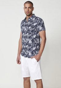 Kratka majica s dugim rukavima v mornarski modri barvi z belim vzorcem listov. V kombinaciji z belimi kratkimi hlačami. Tkanina se zdi lahka in zračna.