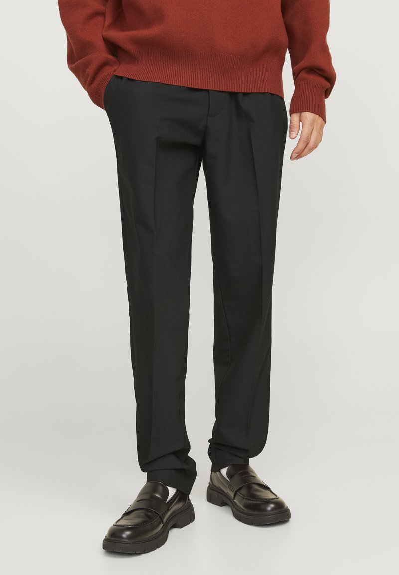 Pantalon noir ajusté en tissu lisse avec une coupe droite et des plis subtils, associé à des mocassins en cuir noir et un pull couleur rouille.