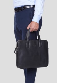 Schwarze Lederaktentasche mit zwei Griffen, einem Reißverschluss und einer glatten Oberfläche. In der Hand einer Person, die blaue Hosen und ein helles Hemd trägt.