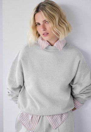 Blonde Frau mit welligem Haar trägt einen hellgrauen Pullover über einem rosa gestreiften Hemd mit Kragen vor einem schlichten hellen Hintergrund.
