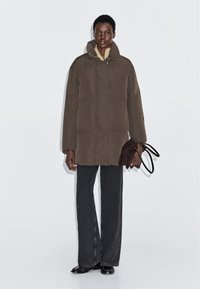 Brun pufferjakke med hætte, der har en afslappet pasform og glat tekstur. Sat sammen med charcoal-stribede bukser og en mørkebrun håndtaske.