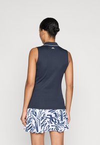 Camisola sem mangas em azul-marinho com textura canelada, apresentando um colarinho branco e um logótipo nas costas, combinada com uma saia branca pregueada com padrões abstractos em azul-marinho.