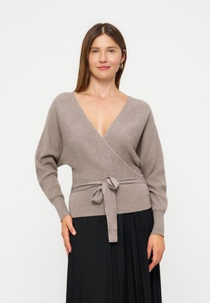 Femme portant un pull cache-cœur taupe avec une ceinture à la taille et une jupe noire plissée, debout devant un fond clair uni.