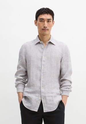 Mężczyzna noszący beżowo-białą, pionowo prążkowaną koszulę z długim rękawem i guzikami, stojący z rękami w kieszeniach na jednolitym tle.