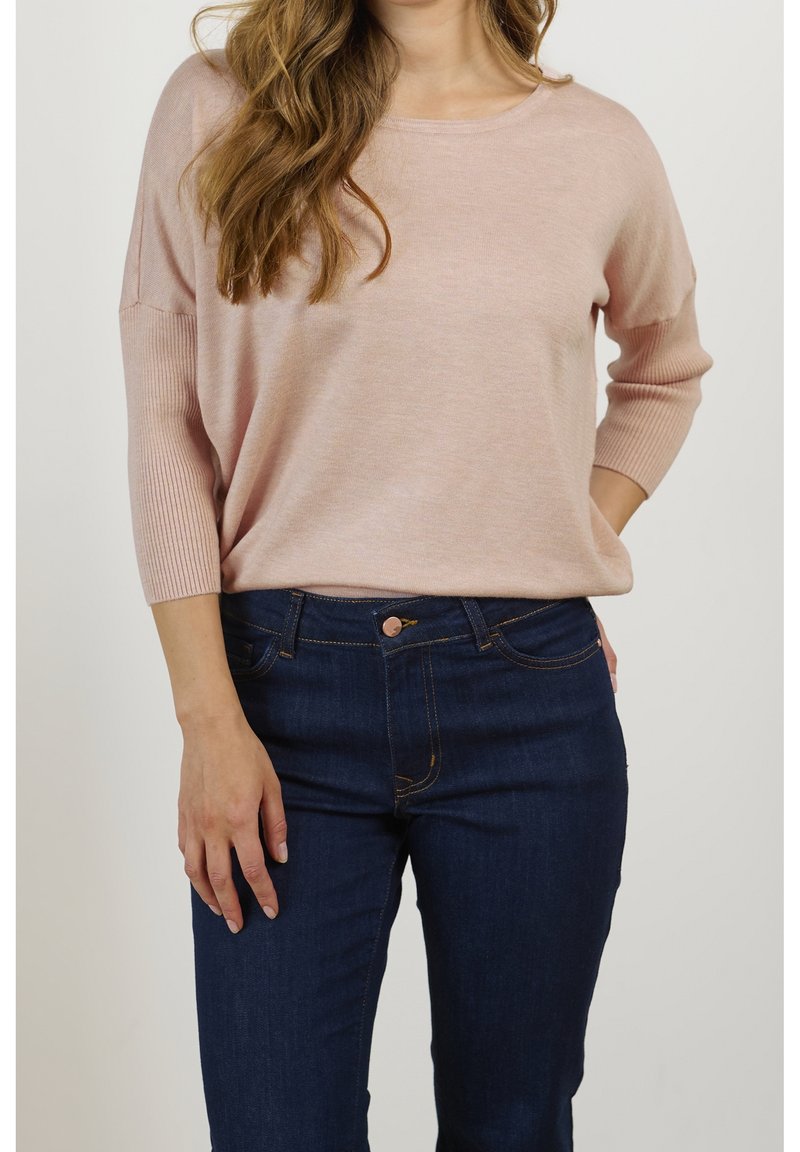 Femme portant un pull rose clair à manches longues et un jean bleu foncé, la main reposant sur la hanche, sur fond uni.