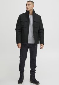 Chaqueta negra aislante con cuello alto, dos bolsillos en el pecho, usada sobre un sudadera gris. Combinada con pantalones oscuros y botas negras de cordones.