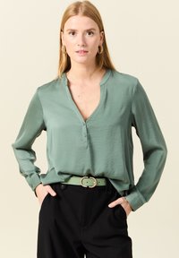 Groene blouse met lange mouwen en een V-hals, met een knopenlijst, gecombineerd met een zwarte riem met een ronde, versierde gesp.