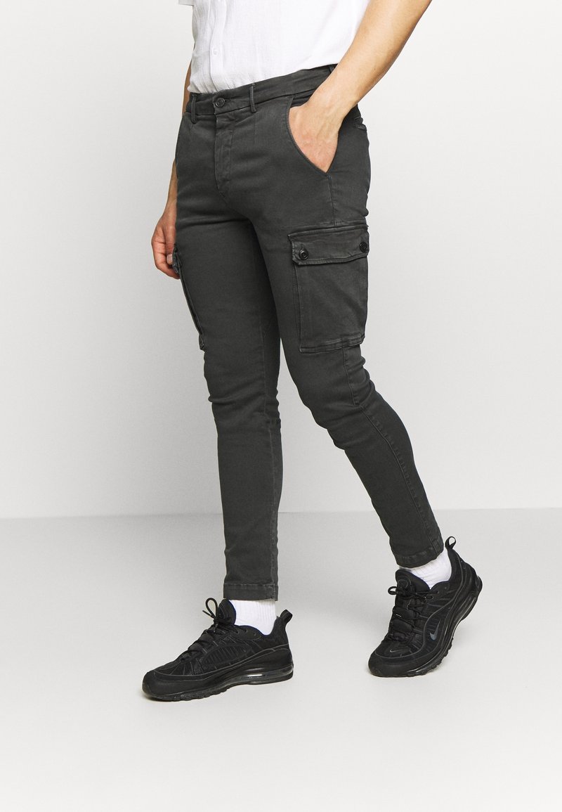 Personne portant un pantalon cargo noir, des baskets noires avec des chaussettes blanches et une chemise blanche, debout avec une main dans la poche.