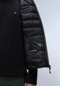 Svart puffjacka med kviltad textur, grön innerficka och logodetalj. Bärs över en svart långärmad skjorta.