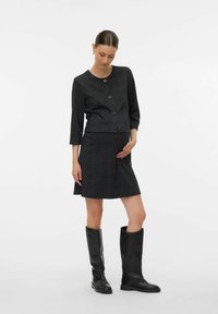 Vrouw in een donkergrijze geruite jas en rok met zwarte kniehoge laarzen, die voorzichtig haar buik vasthoudt, staande voor een effen witte achtergrond.