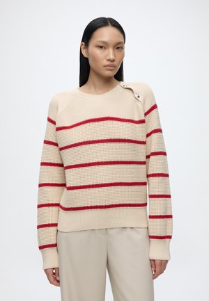 Frau mit glatten schwarzen Haaren, die einen beigen Strickpullover mit horizontalen roten Streifen und beige Hosen trägt, steht vor einem schlichten Hintergrund.