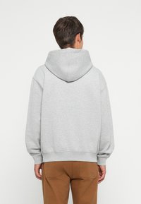 Sudadera gris con bolsillo tipo canguro, corte oversize y dobladillo acanalado. La vista trasera muestra una gran capucha y una tela suave y texturizada. Combinada con pantalones marrones.
