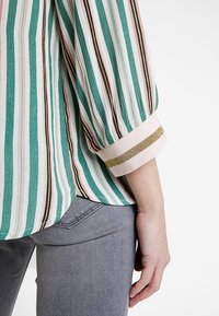 Blusa a righe con colori turchese, rosa, marrone e avorio; tessuto testurizzato con polsini rosa e dettagli dorati.