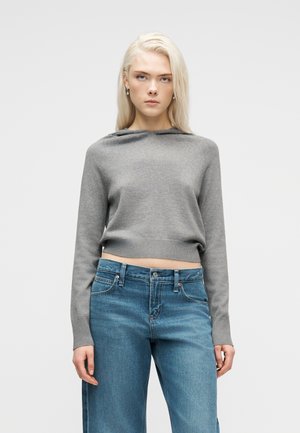 Ξανθιά γυναίκα με γκρι cropped πουλόβερ μακρυμάνικο και ψηλόμεσο μπλε τζιν, στέκεται μπροστά σε απλό φωτεινό φόντο.