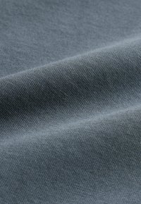 Gros plan sur un tissu doux bleu-gris avec une texture fine en tricot et des plis délicats créant des ombres et des reflets subtils.