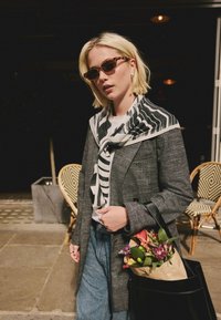 Schwarzer und grauer strukturierter Blazer, weißes T-Shirt, schwarz-weiß gestreifter Schal, Jeans, schwarze Tasche mit Blumen und gemusterte Stühle im Hintergrund.
