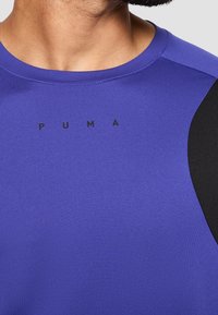 Camiseta deportiva morada con acentos negros, textura suave y cuello redondo. Presenta el logo negro de "PUMA" en el frente.