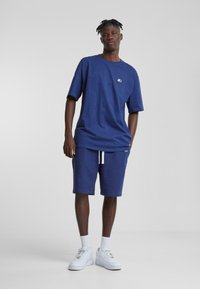 Navy blue short-sleeve t-shirt with small logo, paired with matching shorts featuring white drawstring. Worn with white sneakers.