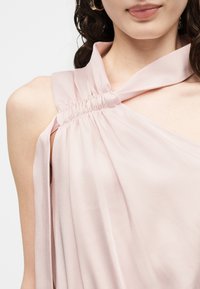 Žena u blijedo ružičastom topu bez rukava s naboranim izrezom i zlatnim naušnicama u obliku obruča, blagi osmijeh na licu, neutralna pozadina.