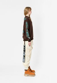 Sudadera marrón con gráficos turquesa, pantalones cargo beige con bolsillos y rayas, y botas de encaje color caqui con suela gruesa.