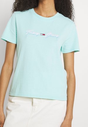 Kobieta ubrana w jasnoniebieski T-shirt Tommy Jeans z logo i białe spodnie, stoi na jednolitym jasnym tle.