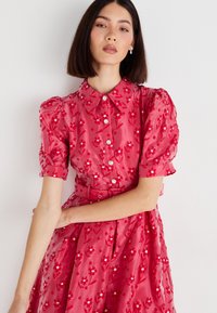 Mujer con cabello lacio y oscuro, viste un vestido rojo con flores, mangas cortas abullonadas, botones de strass y un cinturón a juego, de pie contra una pared blanca.