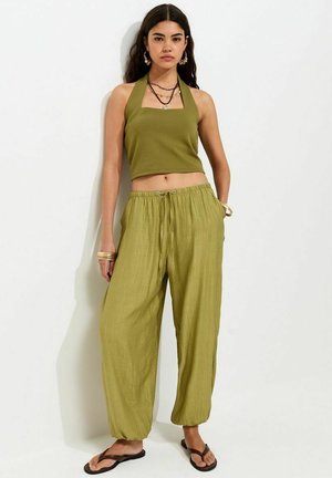 Donna in piedi contro un muro bianco che indossa un crop top verde oliva con collo halter, pantaloni larghi abbinati, infradito nere e gioielli d'oro.