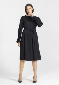 Czarna, długa sukienka z długim rękawem, okrągłym dekoltem, paskiem w talii i teksturowanymi mankietami. Wykonana z gładkiego materiału, długości do kolan, w połączeniu z obcasami.