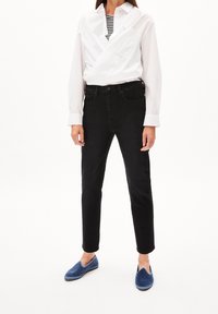 Femme portant une chemise blanche portefeuille sur un top rayé, un jean noir slim-fit et des chaussures bleues sans lacets sur fond blanc.