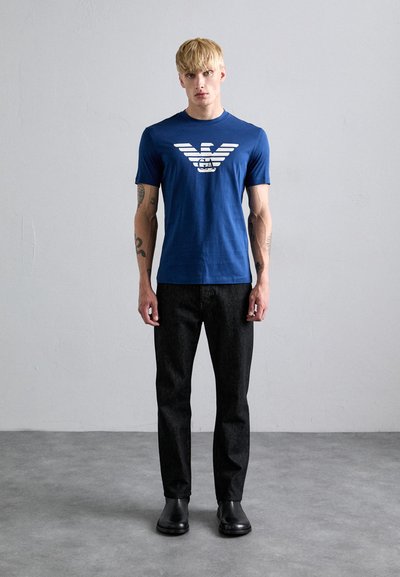 Camiseta de algodón azul con un gráfico blanco con rayas y un emblema de un águila, combinada con pantalones de mezclilla oscuros y zapatos negros.