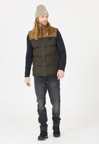 Men's padded vest in dark green and brown, black long-sleeve shirt, gray jeans, and black boots. Worn with a light gray beanie.