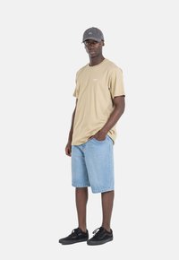 Camiseta de algodón beige con un pequeño logo, pantalones cortos de mezclilla azul claro y zapatillas negras; modelo lleva una gorra gris oscuro. Diseño simple y casual.