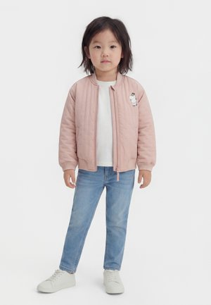 Ungt barn stående vendt fremad iført en lyserød lynlåstrøje med tegneserie-logo, hvid t-shirt, blå jeans og hvide sneakers.