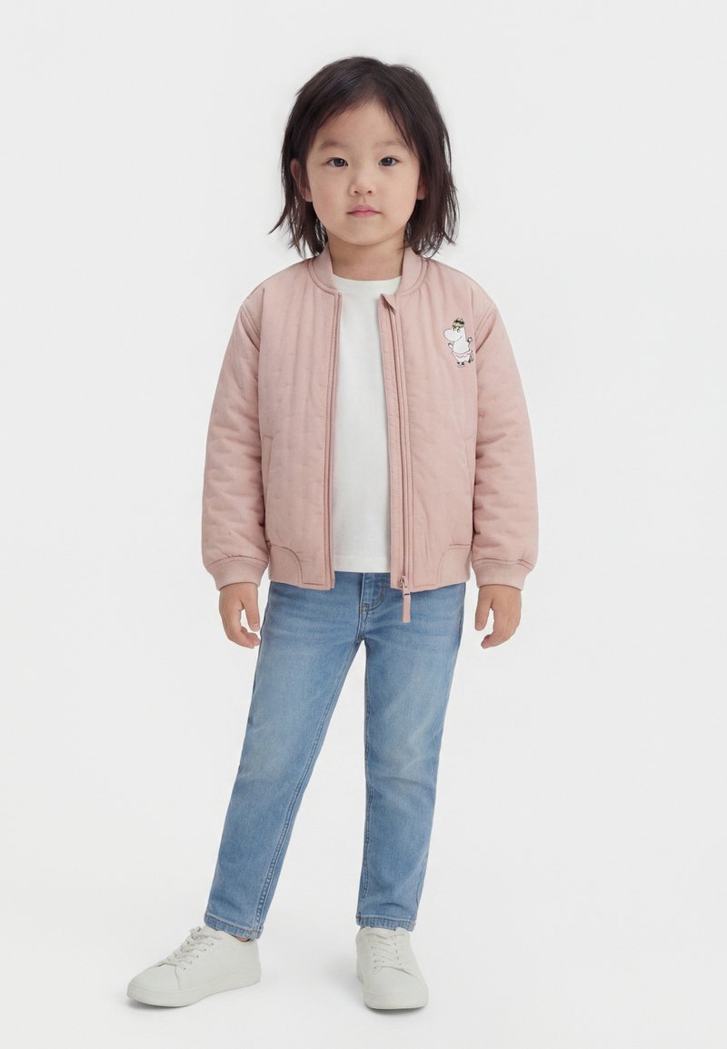 Νεαρό παιδί που στέκεται με το πρόσωπο προς τα εμπρός, φοράει ροζ ζακέτα με φερμουάρ και λογότυπο κινούμενου σχεδίου, λευκό μπλουζάκι, μπλε τζιν παντελόνι και λευκά αθλητικά παπούτσια.