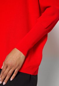 Pull en tricot côtelé rouge avec des manches longues et une coupe décontractée, doté d'une fente sur le côté de l'ourlet et d'une texture lisse.