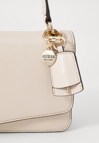 Borsa in ecopelle beige con hardware dorato, caratterizzata da un charm rotondo e dettagli a patta verticale. Texture liscia e cuciture a contrasto.