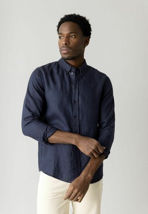 Man met kort krullend haar, gekleed in een marineblauw overhemd met knopen, terwijl hij de mouw oprolt, staand tegen een effen lichte achtergrond.