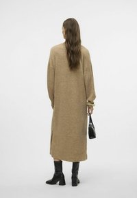 Robe jersey beige longue et ample avec des fentes sur les côtés, des poignets côtelés et une texture lisse, associée à des bottes noires à talons jusqu'aux genoux et à un petit sac noir.