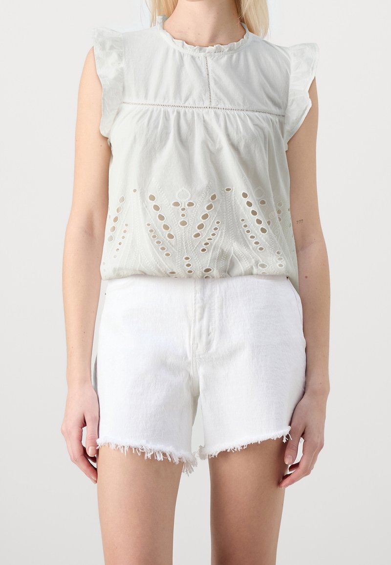 Femme porte un chemisier blanc sans manches avec des détails ajourés et des épaules à volants, assorti d'un short en jean blanc effiloché.